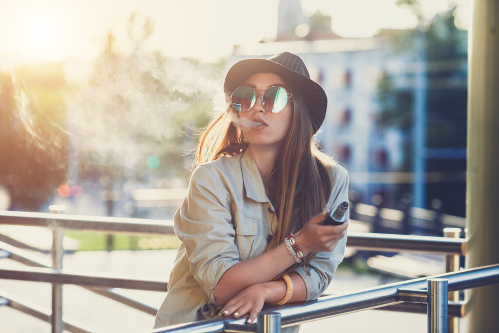 Eine Frau genießt eine entspannte Vaping-Pause im Freien, während sie stilvoll eine Dampfwolke ausstößt. Das Bild symbolisiert modernen Genuss und einen bewussten Lebensstil, unterstützt durch Produkte wie 187 Liquid.